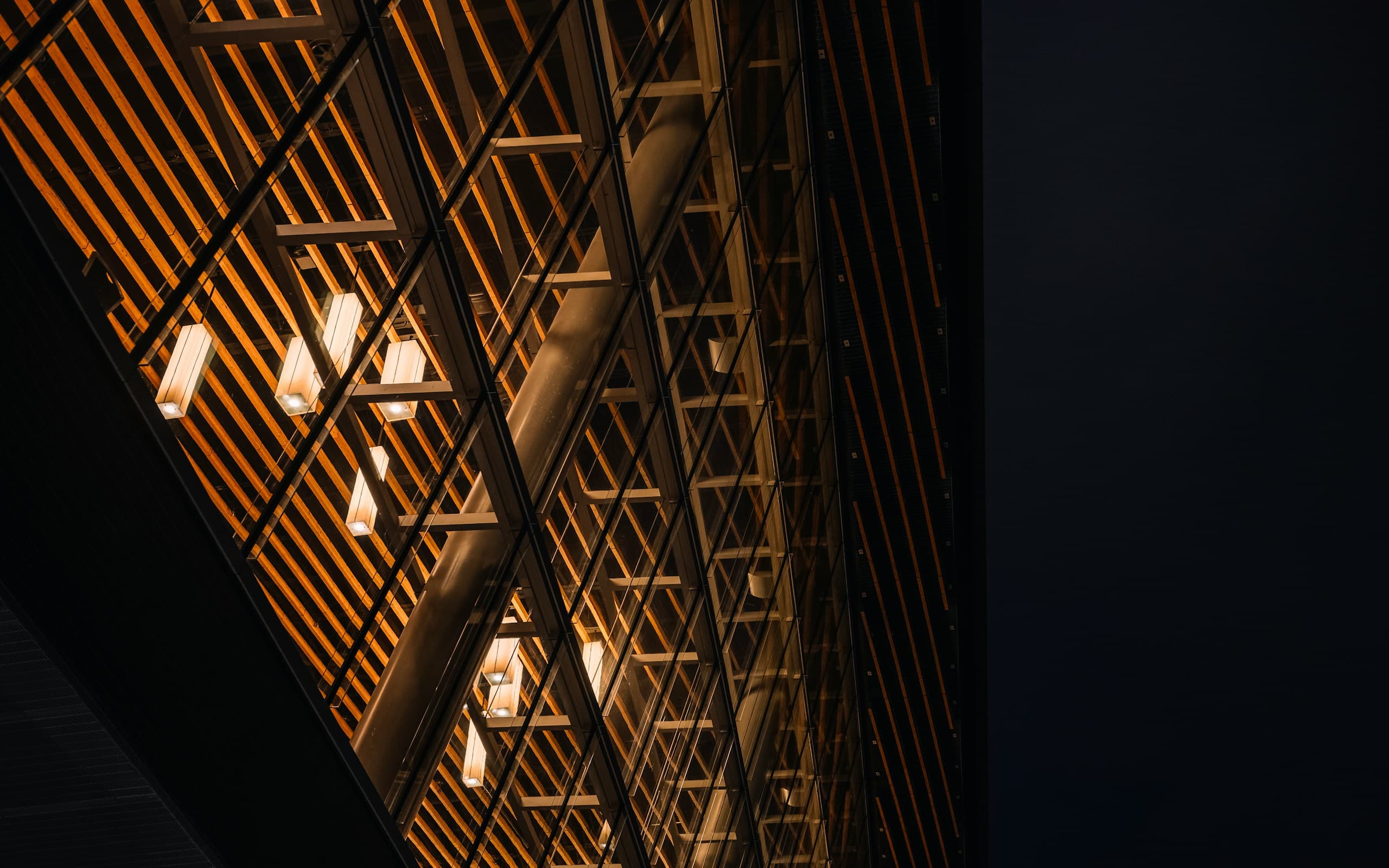 A building facade at night, warm light glowing through a regular grid of timber screens.